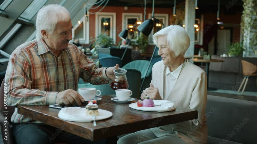 Senior man pouring tea in wife's cup taking care of woman talking and smiling in cafe, delicious desserts are visible on table. People and restaurants concept. Senior man pouring tea in wife's cup taking care of woman talking and smiling in cafe, delicious desserts are visible on table. People and restaurants concept.