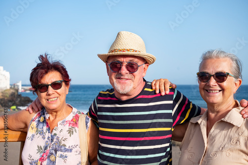 A group of three carefree elderly people looking at camera enjoying the seaside excursion standing in a wooden footpath. Active lifestyle for three retirees