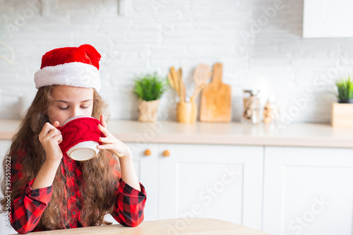 charming happy kid in a New Year's hat sits on the couch and smiles at the camera. girl at home dreams and drinks tea. Family time, love and christmas concept