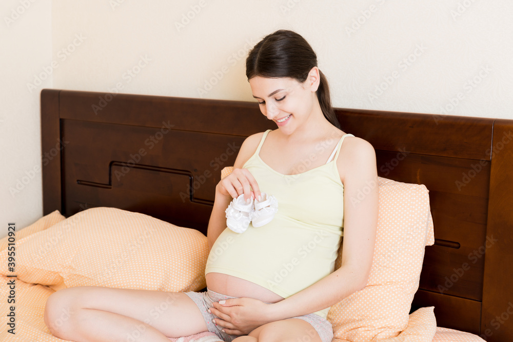 Pregnant woman With Little Baby Shoes sitting on bed in home Pregnant woman With Little Baby Shoes sitting on bed in home