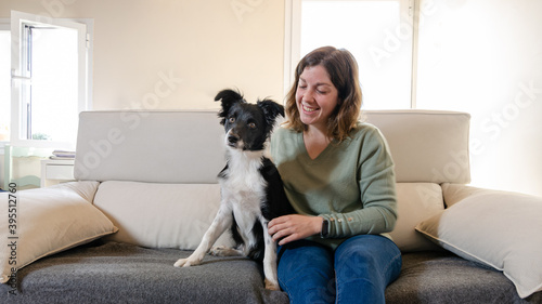 Smiling young woman sitting on the couch hugging a happy border collie puppy. Dog sitting next to young woman