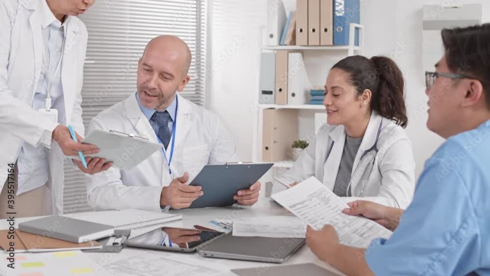 Over shoulder of unrecognizable person sitting at table with male Caucasian and female Mixed-Race colleagues working with papers, talking, cropped doctor leaning down with clipboard for signature Over shoulder of unrecognizable person sitting at table with male Caucasian and female Mixed-Race colleagues working with papers, talking, cropped doctor leaning down with clipboard for signature
