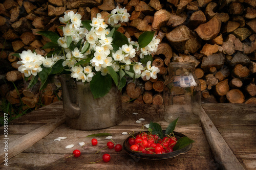 Composition from vintage items on the background of firewood. Jasmine flowers in a teapot, a glass jar and cherry berries are located on a wooden table. Selective sharpness, background blur.