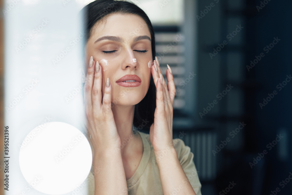 Pretty female with natural makeup applying cream on her face. Brunette young woman using moisturizing cream. Pretty female with natural makeup applying cream on her face. Brunette young woman using moisturizing cream.