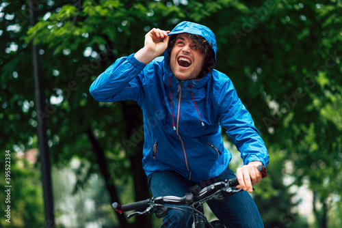 Joyful young man standing with his bike before bicycling on a rainy day next to the house. Happy curly male courier in blue raincoat delivers parcel cycling with a bicycle.