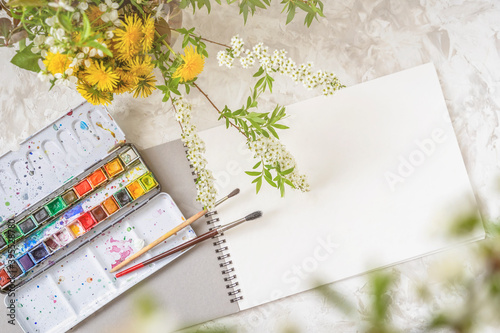 Fototapeta Naklejka Na Ścianę i Meble -  Top view of a table with a sketchbook, paints and brushes next to a bouquet of spring flowers