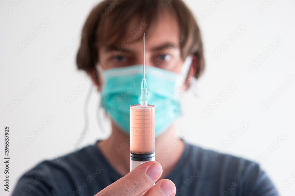 Out of focus Caucasian male wearing a medical mask holding a syringe with orange serum in front of his face