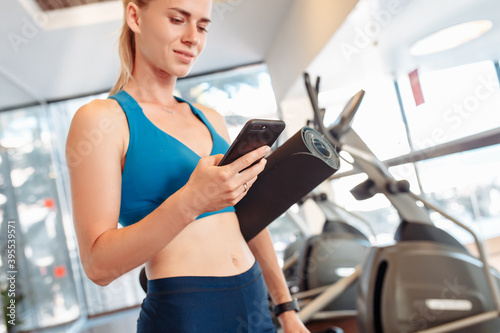 Smiling young woman fitness trainer with exercise mat in hand posing on the background of exercise equipment in the gym. Healthy lifestyle and physical activity concept