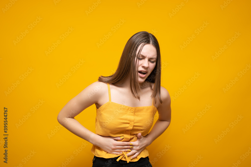 Obraz premium Young beautiful caucasian woman in a yellow tank top on a yellow background with a hand on her stomach because of nausea, painful discomfort. Pain concept.