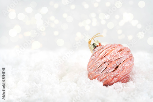 A shiny rose gold Christmas decoration lies on white snow. Glass decoration for a Christmas tree on a white background. Bokeh in the background. Close-up