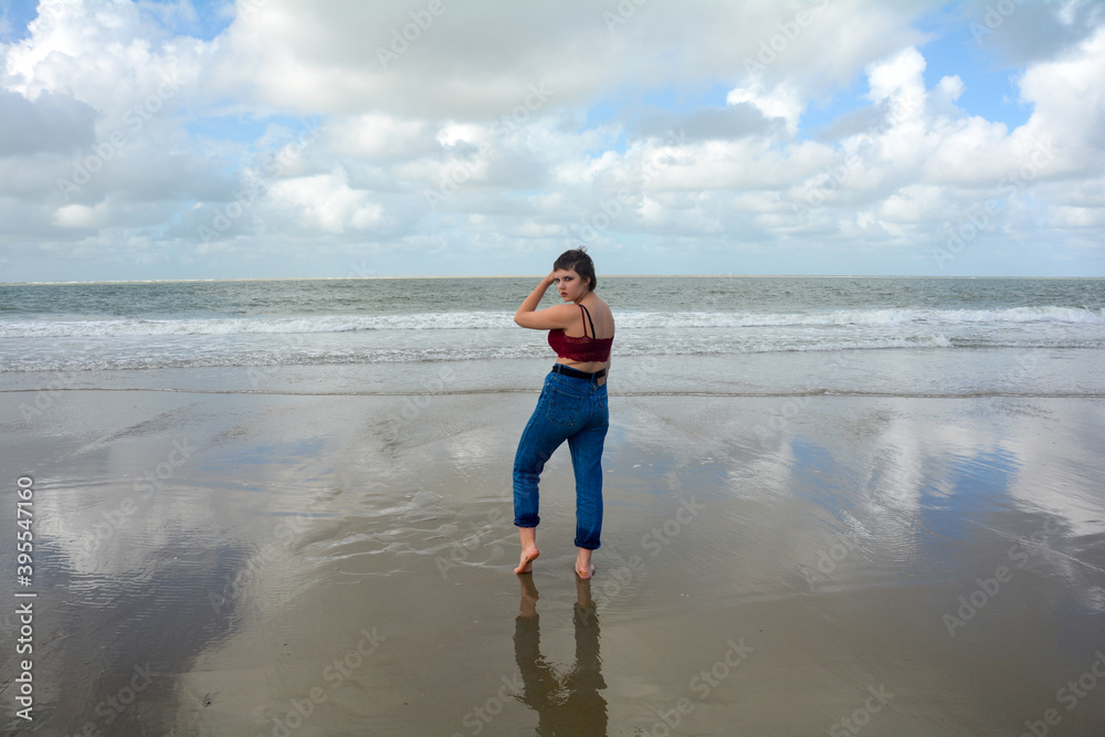 Young woman from the side stands on the beach in front of the sea, arm on head Young woman from the side stands on the beach in front of the sea, arm on head