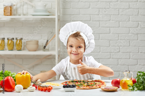 little boy in chef hat and an apron cooking pizza in the kitchen. child showing Ok gesture. delicious