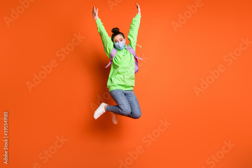 Full length photo portrait of girl raising hands up jumping wearing blue fabric face mask isolated on vivid orange colored background