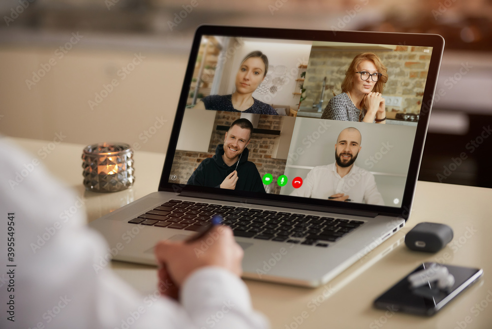 A laptop screen view of the telecommunications application during an online meeting over a man's shoulder. Back view of an employee who is doing notes on a video conference on a computer from home