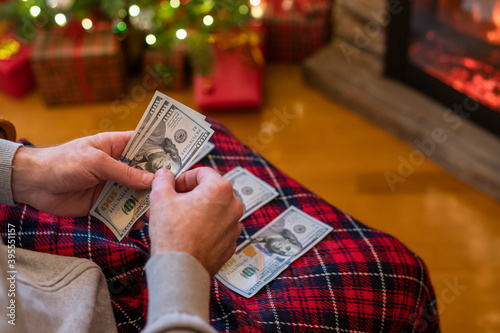 Man counting american dollars sitting near christmas tree and fireplace. Spending money on gift at christmas time. concept.