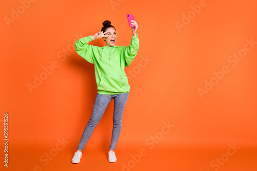 Full length photo portrait of girl making v-sign near eye holding phone in one hand laughing taking selfie isolated on vivid orange colored background © deagreez