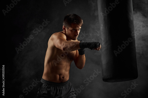 Male boxer training defense and attacks in boxing bag on black background with smoke