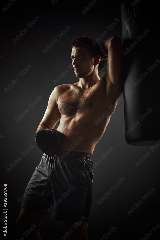 shirtless handsome athlete boxer in black gloves standing near punching bag on dark background