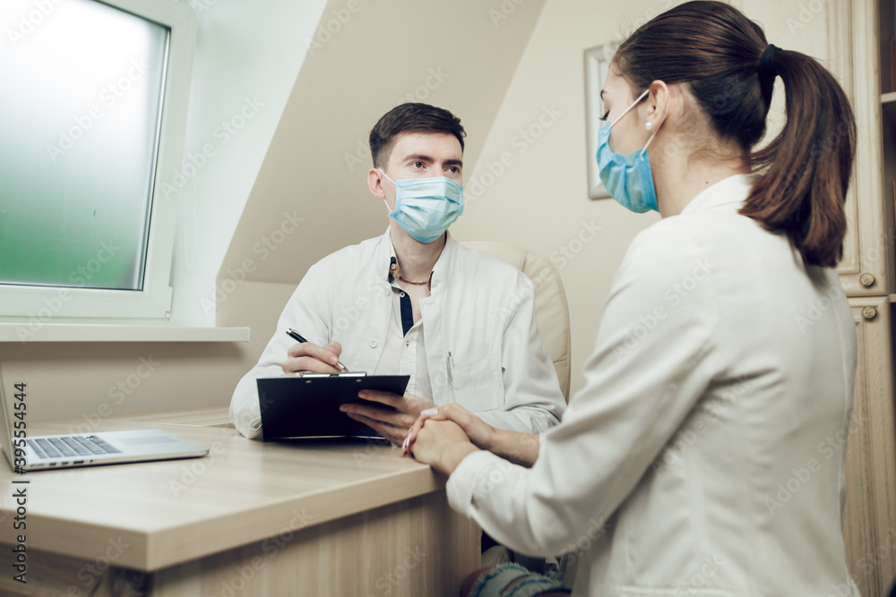 Admission to the doctor during quarantine. Young doctor and patient sitting in the office wearing protective masks. The doctor writes a prescription to a young female patient Admission to the doctor during quarantine. Young doctor and patient sitting in the office wearing protective masks. The doctor writes a prescription to a young female patient