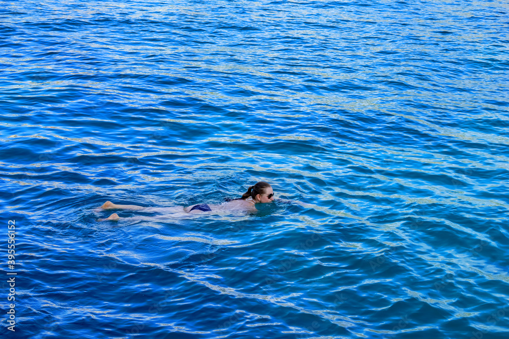Adult girl in bikini and sunglasses lies on the surface of the water. Young woman swimming in blue sea water - top side view