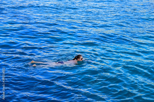 Adult girl in bikini and sunglasses lies on the surface of the water. Young woman swimming in blue sea water - top side view