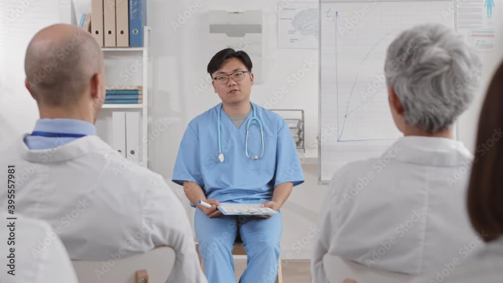 Over shoulder medium long of young Asian male doctor wearing blue scrubs, stethoscope on neck, glasses, sitting with clipboard in hands, making report to unrecognizable colleagues Over shoulder medium long of young Asian male doctor wearing blue scrubs, stethoscope on neck, glasses, sitting with clipboard in hands, making report to unrecognizable colleagues