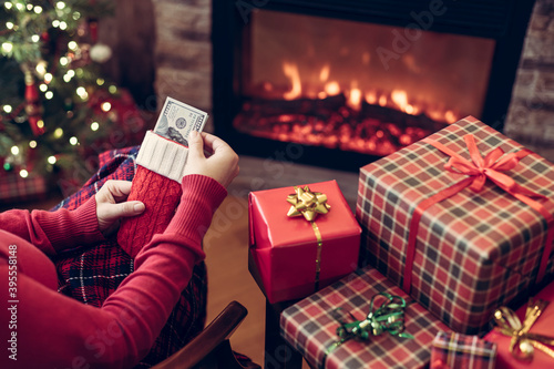 Woman putting american dollars in red christmas sock sitting near christmas tree and fireplace and packing gift box. concept.