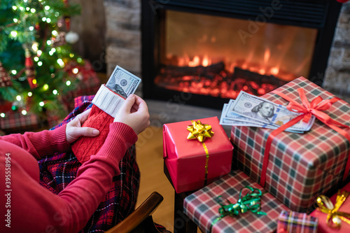 Woman putting american dollars in red christmas sock sitting near christmas tree and fireplace and packing gift box. concept.