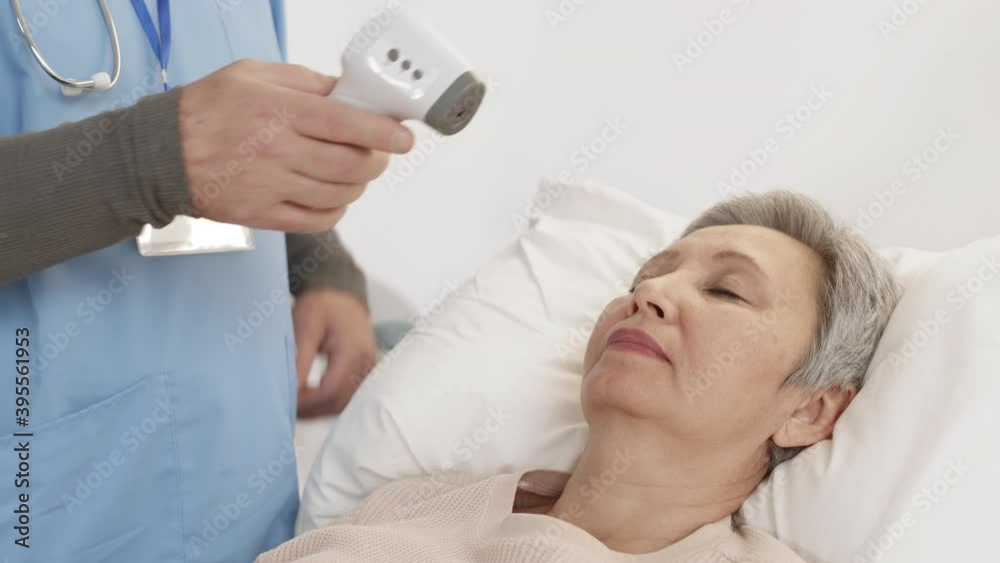 Close up of elderly Asian woman sleeping in bed in hospital ward while unrecognizable doctor wearing blue uniform checking her temperature using non-contact thermometer Close up of elderly Asian woman sleeping in bed in hospital ward while unrecognizable doctor wearing blue uniform checking her temperature using non-contact thermometer