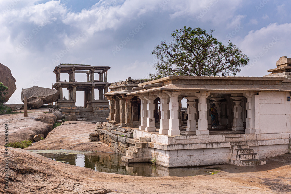 Hampi, Karnataka, India - November 4, 2013: Virupaksha Temple complex. Ruined Moola white pillar section on hill under blue cloudscape with small pond, and double story 