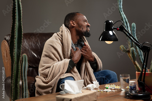Fever. Young African-american man wrapped in a plaid looks sick, ill sitting on armchair at home indoors. Healthcare and medicine, ill prevention, seasonal illness symptoms and self-protection.