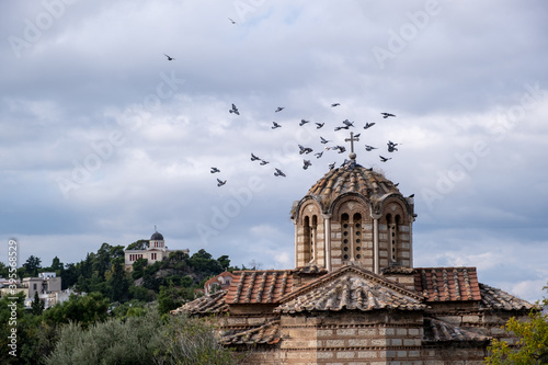 Greek Orthodox church in Thissio area, Athens, Greece. Flock of pigeons flying around the dome, cloudy sky background