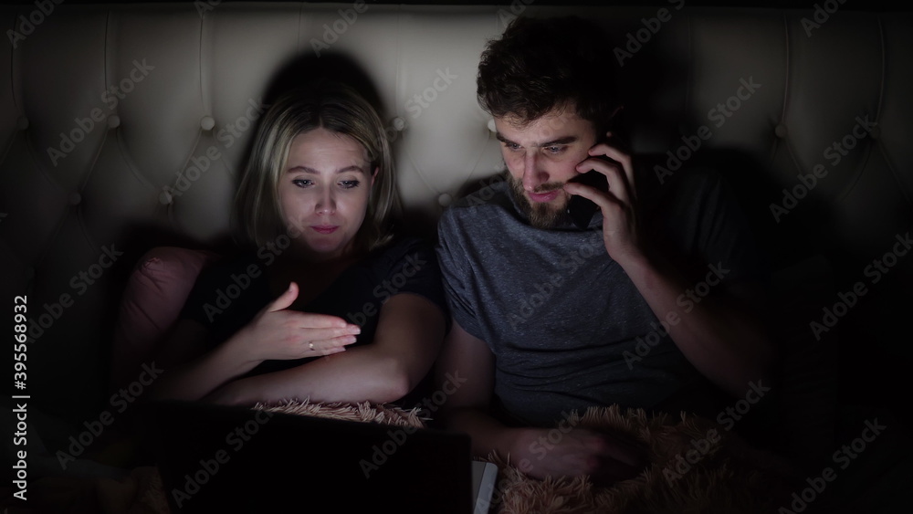 A man and a woman, a young couple are watching a movie on a laptop in bed before going to bed and they are attracted by a mobile phone call