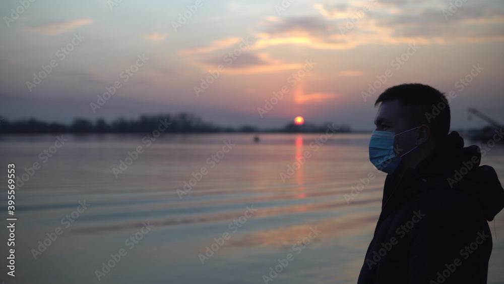 A man in a mask stands by the river, lake and looks at the sunset