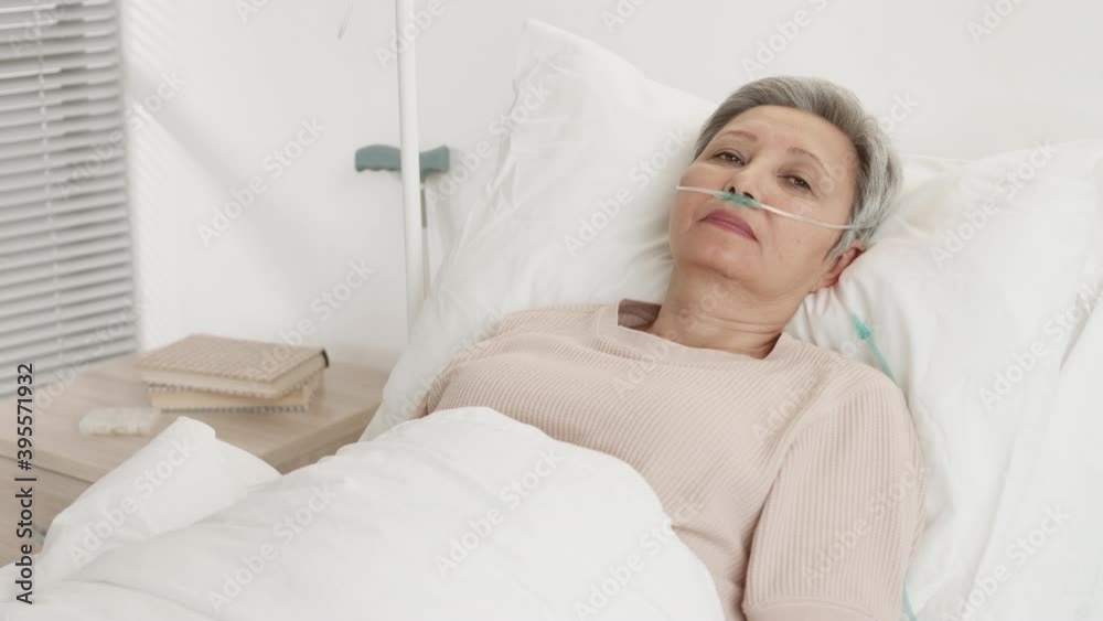 Medium shot of ill senior Asian woman with short hair and oxygen tube in her nose lying in bed in hospital room and looking at camera
