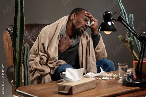 Terrible headache. African-american man wrapped in a plaid looks sick, ill sitting on armchair at home indoors. Healthcare and medicine, ill prevention, seasonal illness symptoms and self-protection.