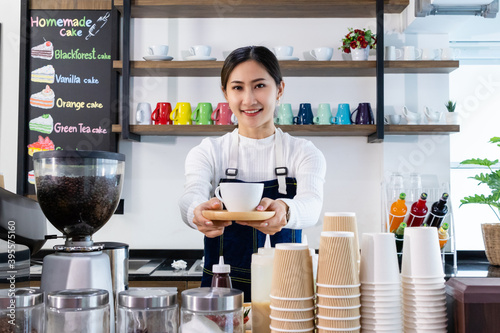 Young Asian woman barista work in coffee shop, holding a cup of coffee behind counter, coffee maker and supplies. Small business owner or worker smile and ready to service.