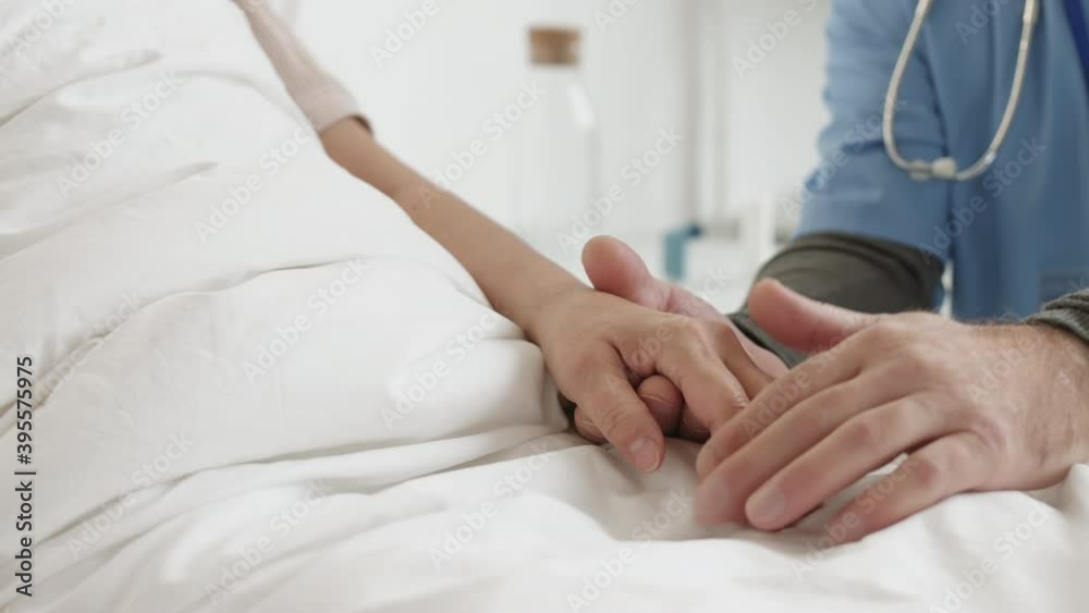 Close up of unrecognizable male doctor sitting near bed in hospital ward and patting unrecognizable female patient hand Close up of unrecognizable male doctor sitting near bed in hospital ward and patting unrecognizable female patient hand