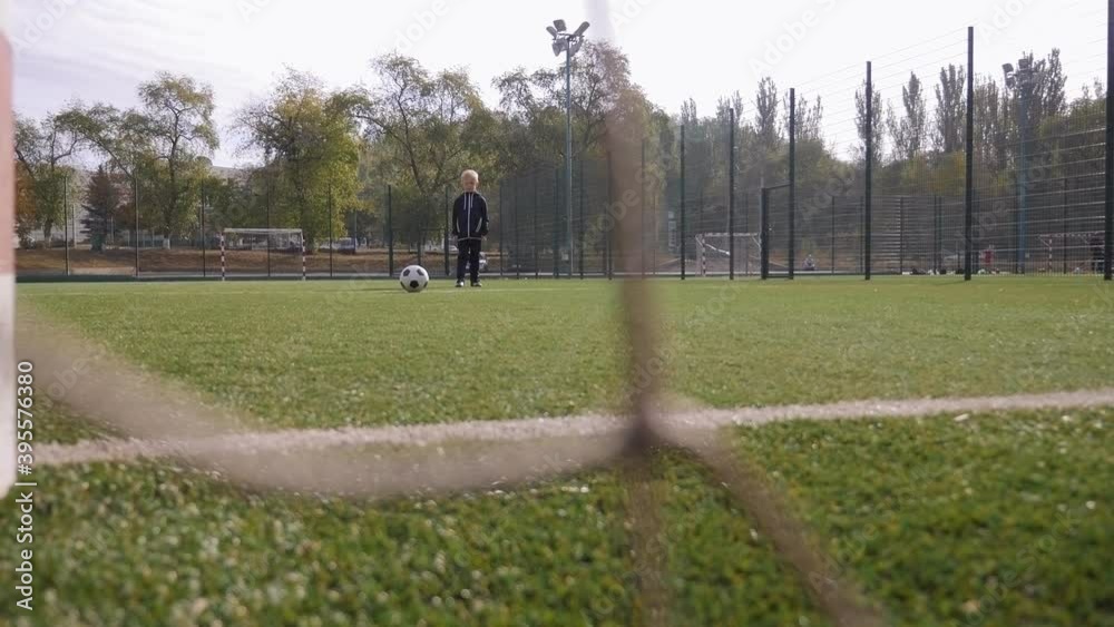 A small boy soccer player hits the goal with a soccer ball, in the foreground a blurred football net. A small boy soccer player hits the goal with a soccer ball, in the foreground a blurred football net.
