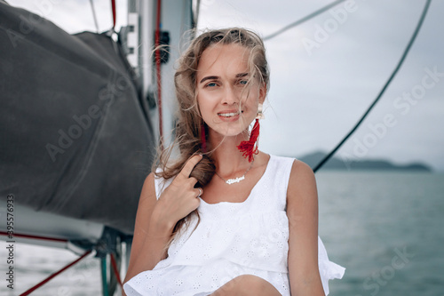 portrait of happy pretty girl with white dress, long red earrings and long curly blonde hair standing on yacht at summertime. looking at camera with toothy smile. close UP