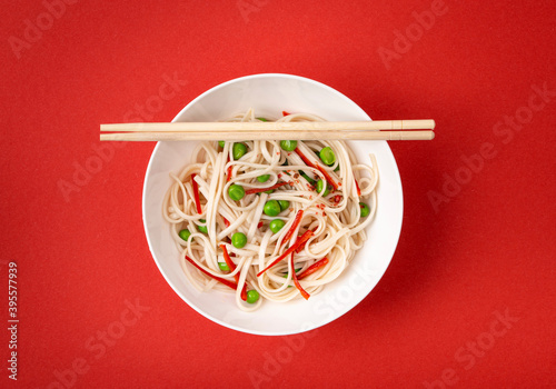 Asian noodles with vegetables, green peas and red pepper in white bowl with wooden chopsticks photo