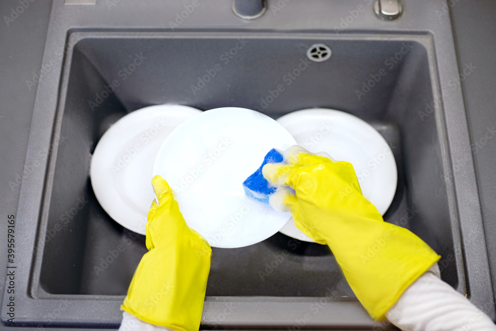 A woman washes white dishes with a blue sponge and yellow gloves in the kitchen sink. A woman washes white dishes with a blue sponge and yellow gloves in the kitchen sink.