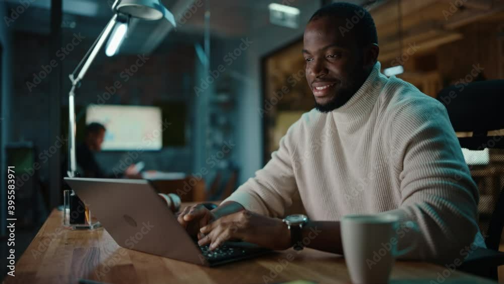 Handsome Black African American Man Having an Online Conversation on a Laptop Computer in Creative Office Environment. Happy Male is Browsing Social Media and Replying to Friends in Messenger. Handsome Black African American Man Having an Online Conversation on a Laptop Computer in Creative Office Environment. Happy Male is Browsing Social Media and Replying to Friends in Messenger.
