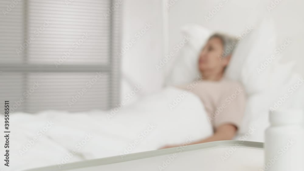 Racked focus shot of unrecognizable male nurse wearing blue overall giving glass of water to sick old Asian woman lying in bed in hospital room Racked focus shot of unrecognizable male nurse wearing blue overall giving glass of water to sick old Asian woman lying in bed in hospital room