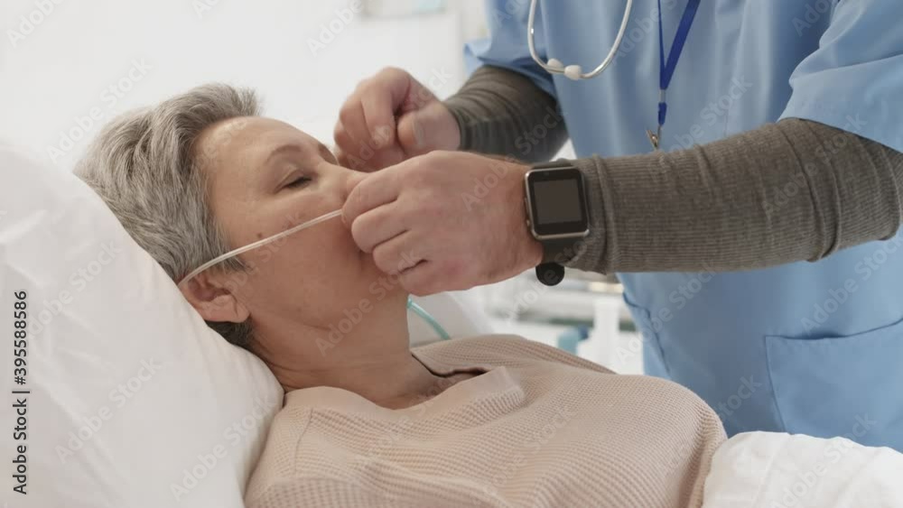 Lockdown of unrecognizable male doctor coming to check on aged Asian woman with short hair and oxygen tube in her nose lying in coma in bed in hospital ward and patting her on head Lockdown of unrecognizable male doctor coming to check on aged Asian woman with short hair and oxygen tube in her nose lying in coma in bed in hospital ward and patting her on head