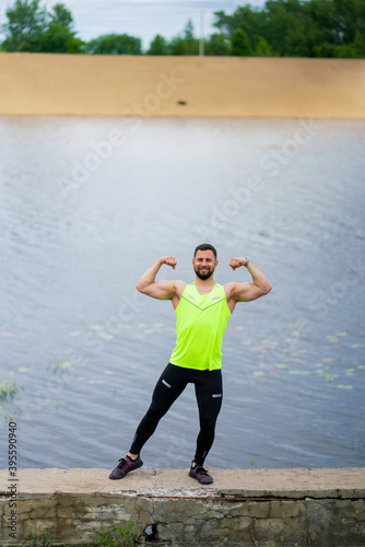 Happy athletic young guy in stylish sportswear posing showing muscles outdoor near water