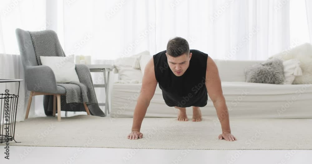 Sporty athletic male exercising on his hard workout on quarantine. Intense training in modern light living room. Young man is doing push-ups exercises at home keeping himself during lockdown.