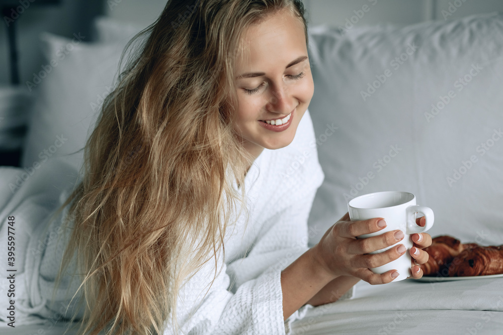 Beautiful blonde woman in white coat with white smile having breakfast in a hotel room. Woman is having breakfast with croissant in the morning!. Beautiful blonde woman in white coat with white smile having breakfast in a hotel room. Woman is having breakfast with croissant in the morning!.