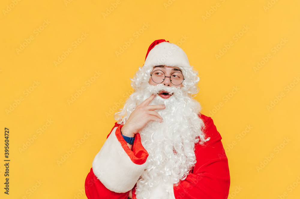Thoughtful Santa Claus is standing at the studio on the yellow background, touching his beard with a thinking gesture, looking aside with his mouth open. Thoughtful Santa Claus is standing at the studio on the yellow background, touching his beard with a thinking gesture, looking aside with his mouth open.