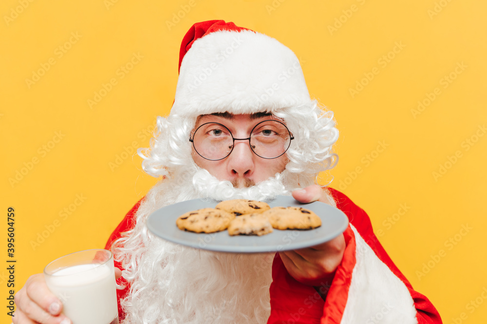 Closeup photo of Santa Claus smelling the tasty chocolate cookies, holding a glass of milk, feeling delighted, standing on an isolated yellow background.
